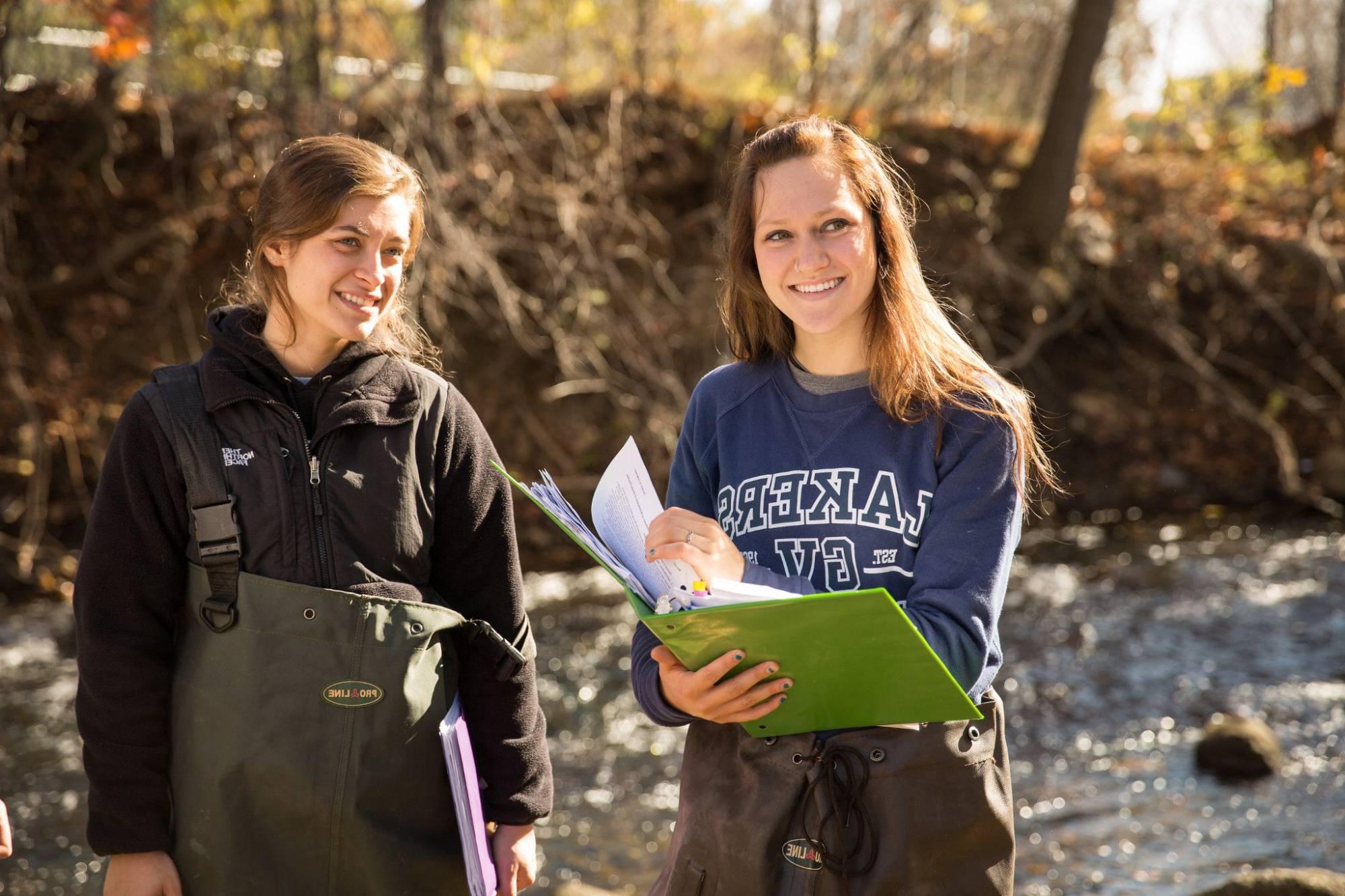 Two young white women on a field experience for a Biology course; they are standing by a river, both are wearing wading gear, and one is holding a large binder.